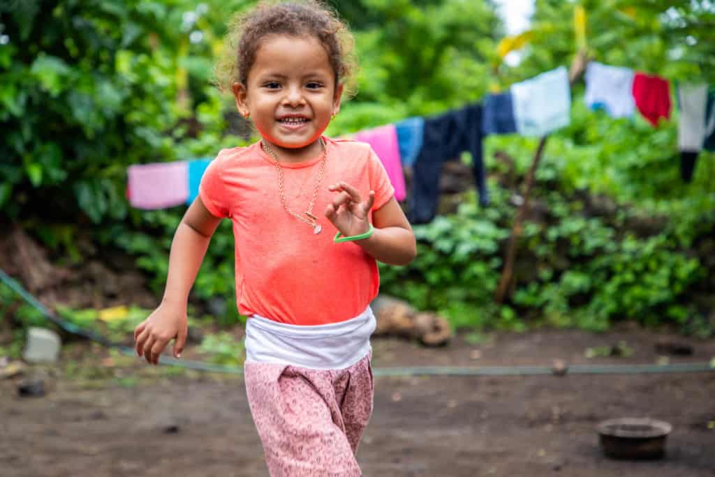 Little girl running outside her house. She is wearing an orange shirt and a pink skirt. Behind her are clothes hanging on a clothesline.