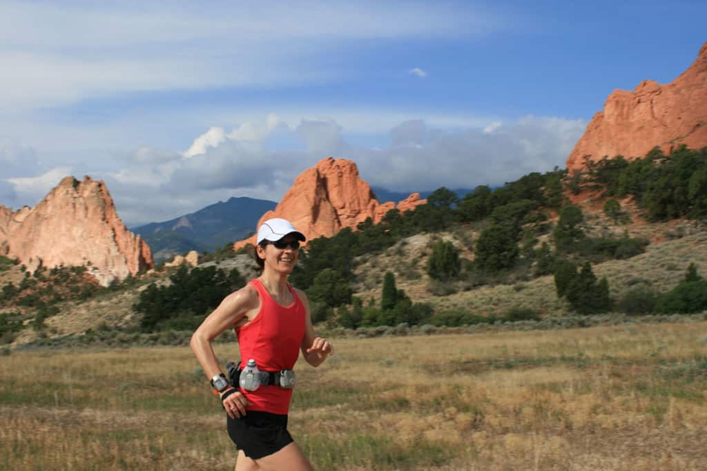 A woman smiles while running in a wilderness area. Behind her are jagged red rocks, blue sky and mountains. She is wearing a white hat, red shirt and black shorts, plus some running accessories.