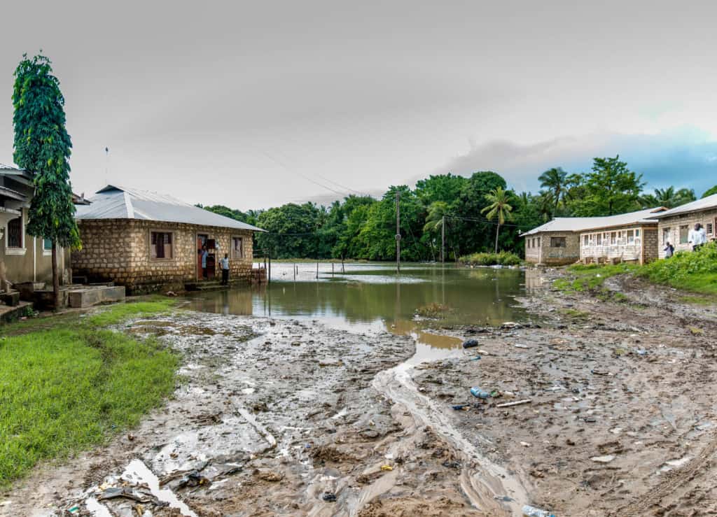 Adults stand outside homes surrounded by floor water, wet mud, grass and trees.
