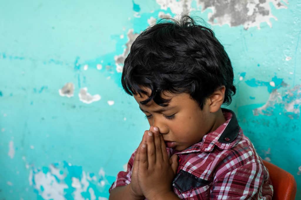 A boy in a red and black plaid shirt sitting in a chair with his eyes closed, and hands clasped together, praying. There is a light blue wall with chipped paint in the background.