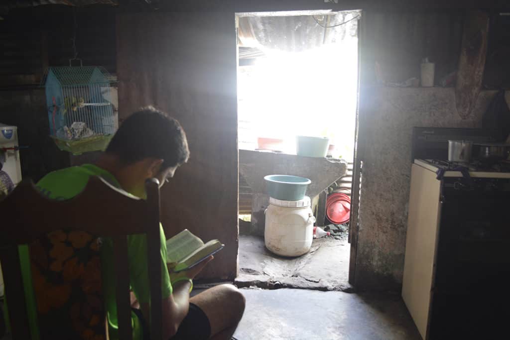 A teenage boy wearing a green shirt and black shorts sits in a wooden chair, reading a Bible. Light streams in through a door.