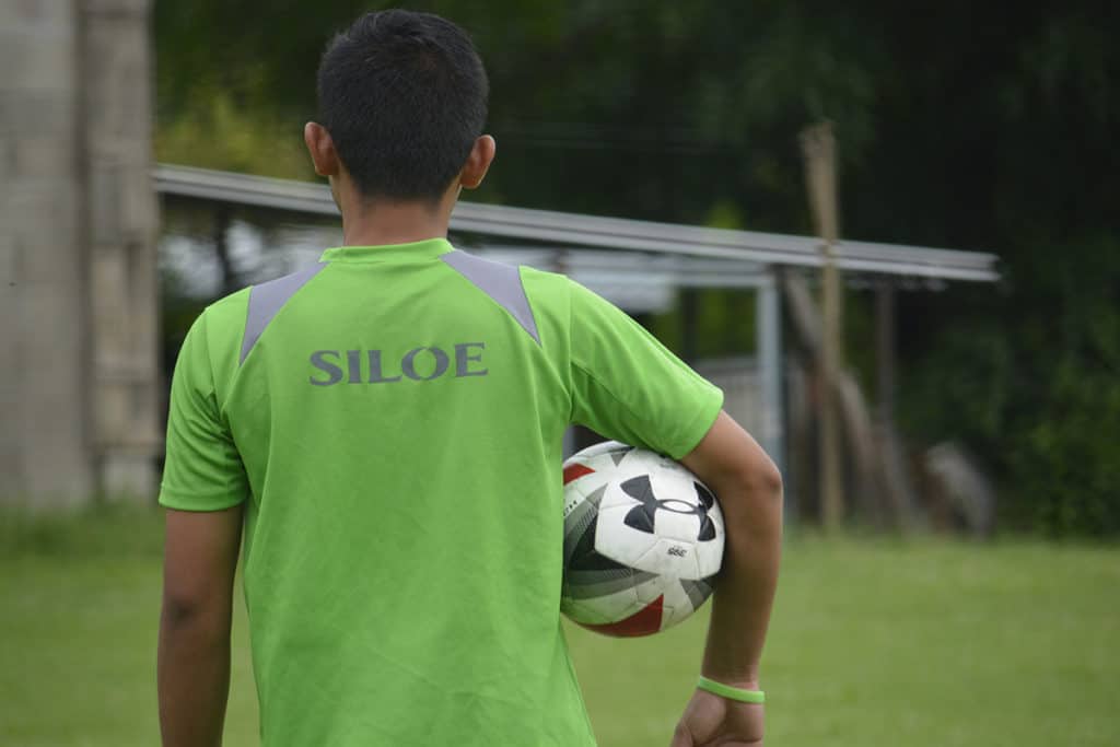 an 18 year old teenager, teen, boy, wearing a green shirt with the word "SILOE" on the back, faces away and under one arm holds a soccer ball, athletic ball, playing a sport, kickball, football, soccer field, in neighborhood.