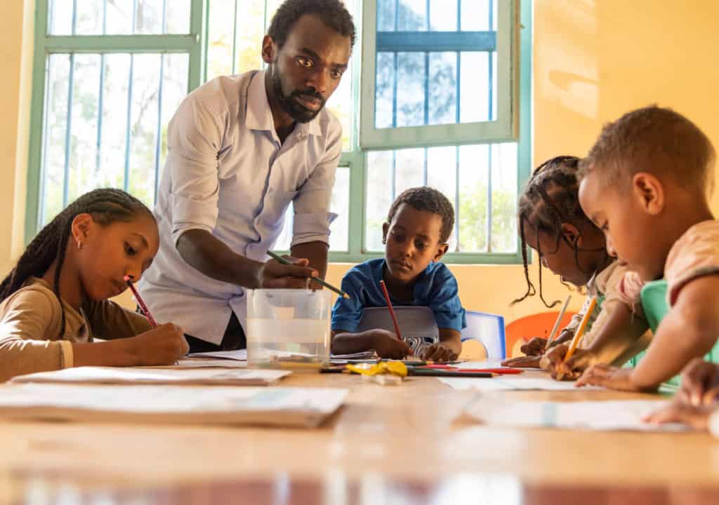 Kenenisa is wearing a blue shirt. He is sitting around a large table in a classroom with yellow walls. He and several other children are writing letters to their sponsors and drawing pictures for them. The children are using colored pencils. The project's social worker, Misgana, wearing a white shirt, is helping the children.