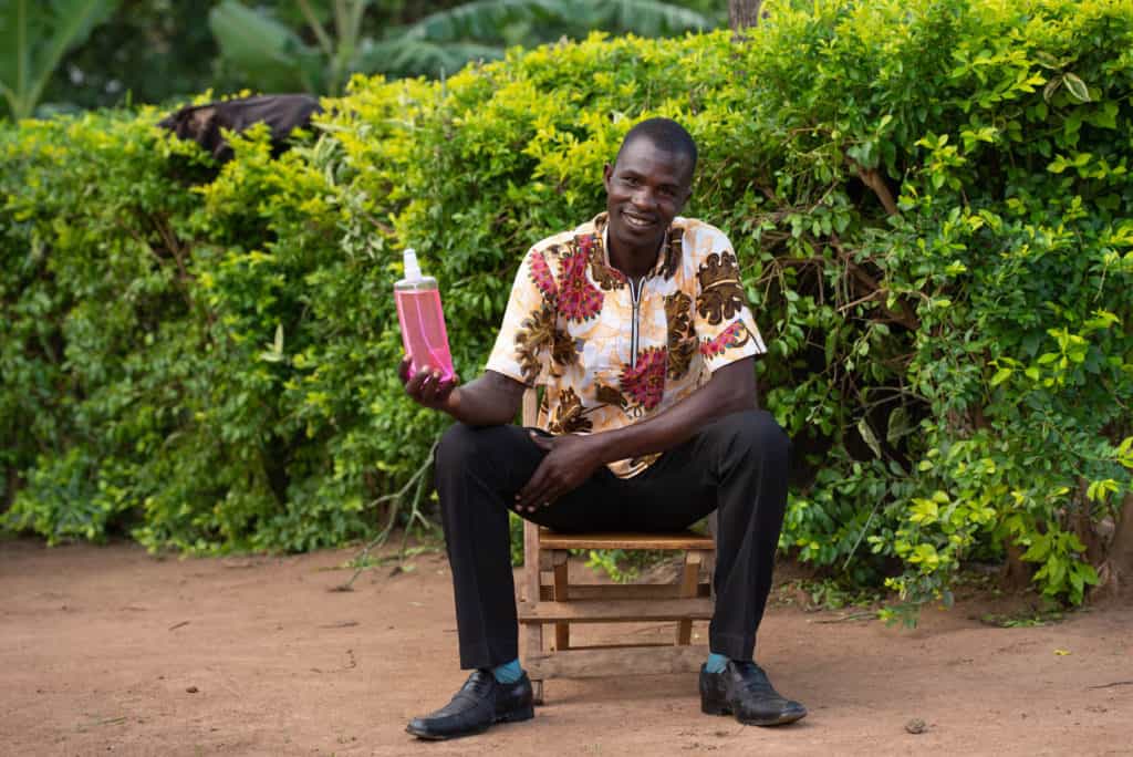 Man wearing black pants and a white shirt with a brown and red floral print. He is sitting on a bench outside his home and is holding up a bottle of hand sanitizer. There are green bushes behind him.
