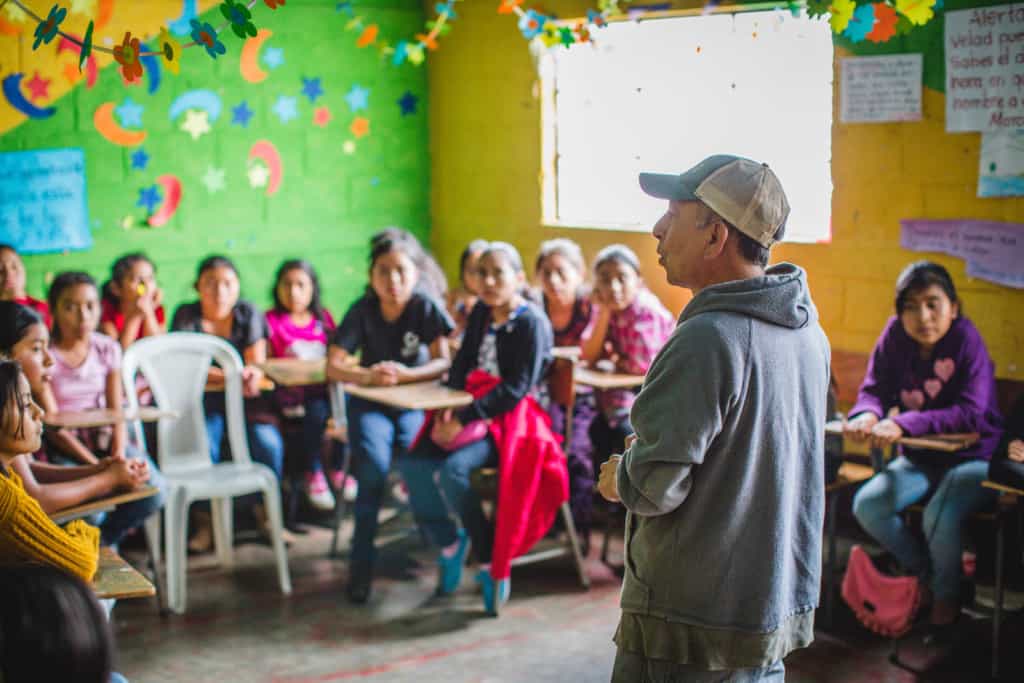 A man in a gray sweatshirt and baseball cap, hat stands in a classroom in front of a group of girls who are sitting at small wood school desks. The walls are yellow and green with a bright window on one wall.