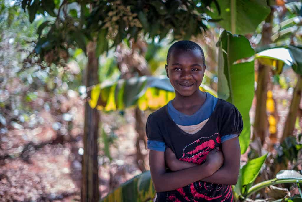 A 12-year-old girl wearing black and red smiles and crosses her arms as she stands outside. There are trees and large green leaves behind her.