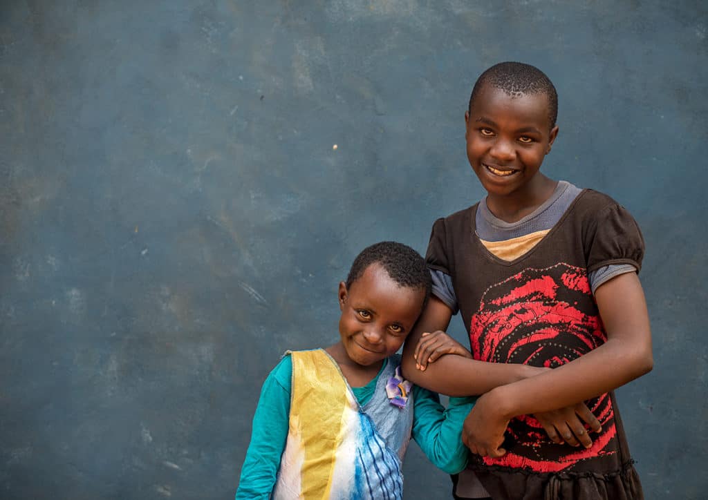 A young girl wraps her arm around the arm of her older sister. The younger sister is wearing a colorful outfit. The older sister is wearing a black and red shirt. Both girls are smiling.