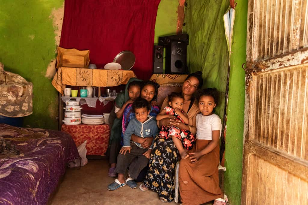 Mother wearing a tan sweater and a black floral skirt. She is sitting in her home surrounded by her children.