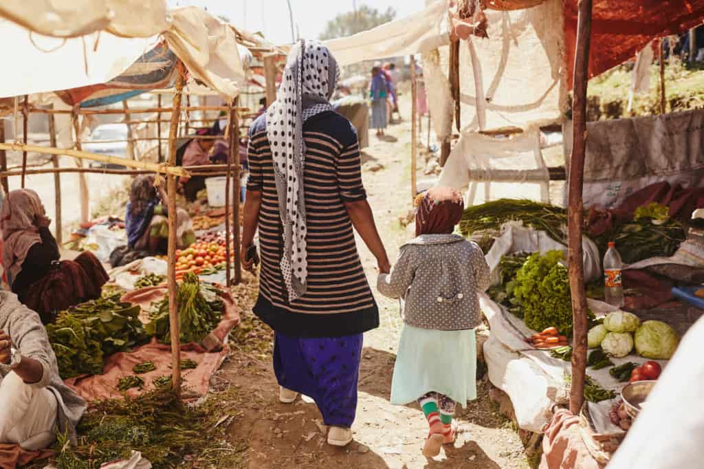 Mother, in a blue skirt, and her daughter, in a brown head covering, hold hands as they walk through the market where the mother makes a living selling vegetables. There are stalls with women sitting on the ground surrounded by produce. The stalls are covered by tarps, fabric.