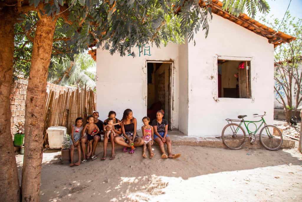 Ana Maria and her family are sitting in front of their home. The house is white. To the right is a green bicycle and there are trees on the left.