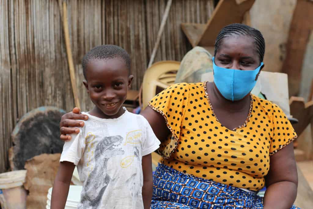 Boy wearing a white shirt with a dinosaur on the front. He is outside his home with his mother, wearing a blue skirt, yellow shirt, and a blue face mask.