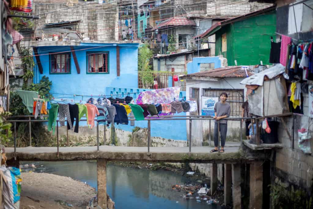 Young man wearing a gray shirt and gray pants. He is standing on a bridge next to a clothesline with clothes on it and he has his hand on a metal railing. Behind him are buildings.