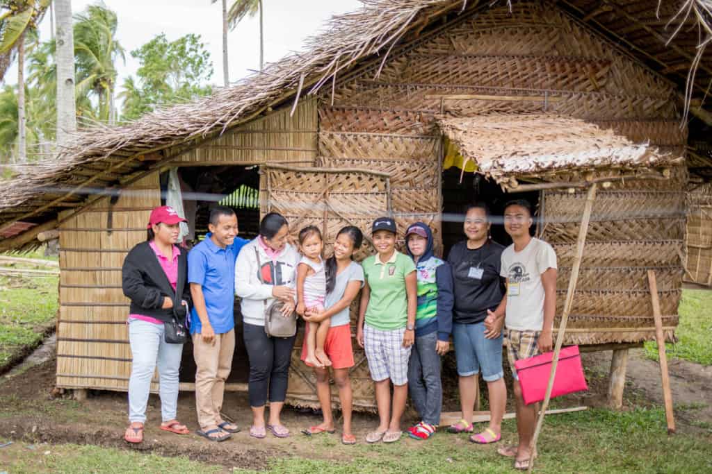 The pastor and staff of a local church during a visit to complete a household survey. They are all standing in front of the home, made of straw.
