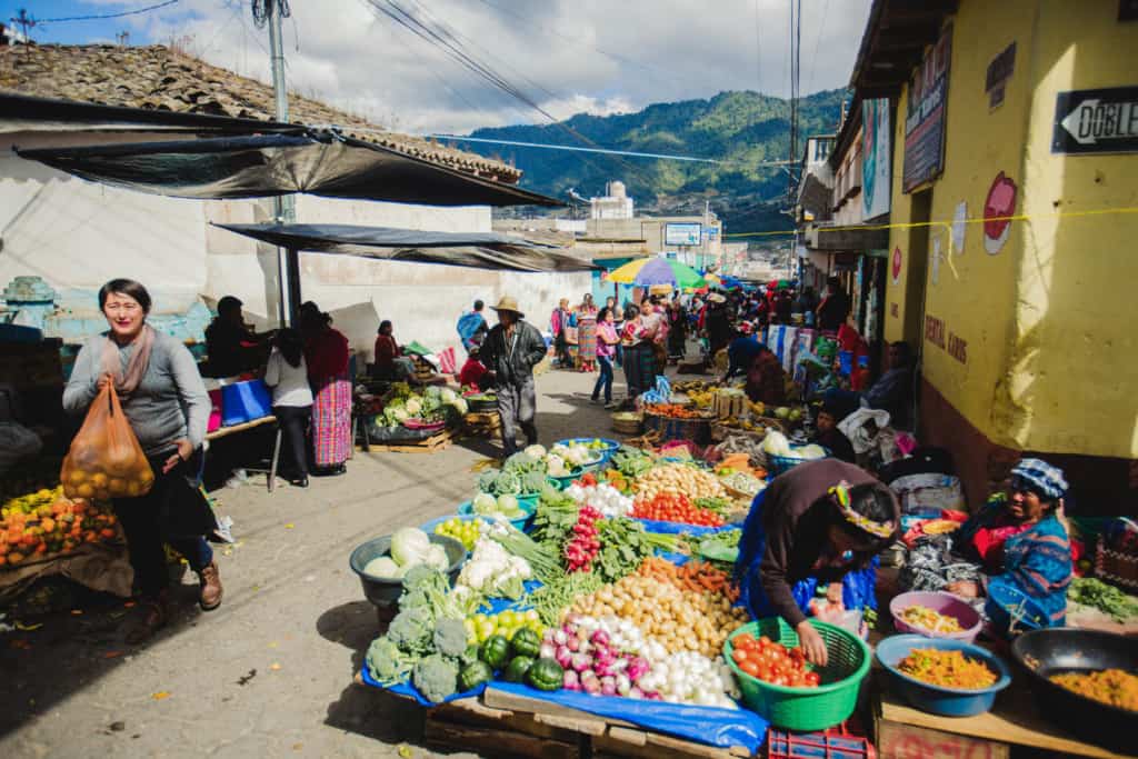 Vegetables are on blue tarps at a street market in Cantel. A woman in a gray shirt walking on the street carries an orange bag full of produce. Another woman leans over a green plastic basket full of tomatoes next to a yellow building. There are hills and more people in the background.