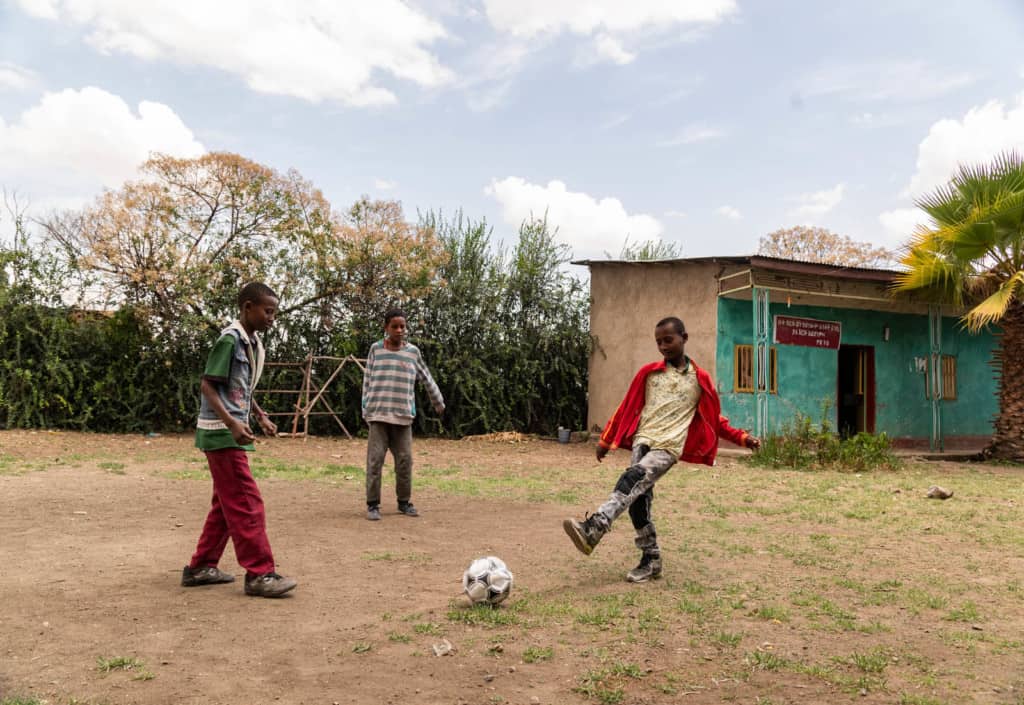 Boy, in a red jacket, outside in the yard at the church kicking around a soccer ball with friends. There is a tan and teal building in the background.