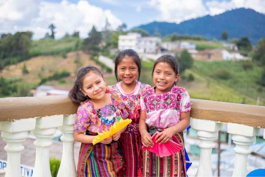 Three girls standing together and smiling, holding yellow and pink handmade paper fans. They are wearing pink shirts and striped skirts. They are leaning against a railing with white pillars. In the background are white buildings and a green hill.