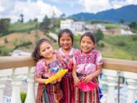 Three girls standing together and smiling, holding yellow and pink handmade paper fans. They are wearing pink shirts and striped skirts. They are leaning against a railing with white pillars. In the background are white buildings and a green hill.