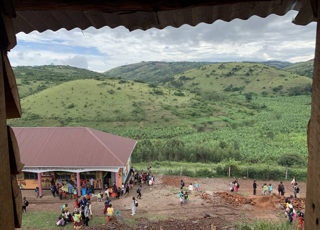 an aerial view of a building surrounded by children, in the background are green hills and a cloudy sky