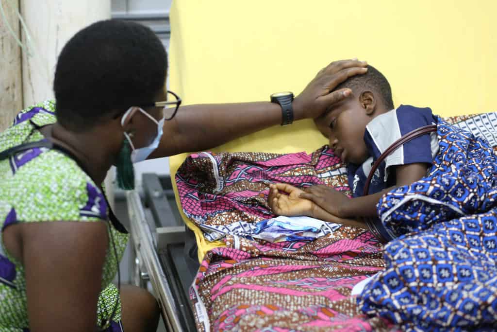 Boy wearing a blue and white shirt and is laying in a hospital bed. A project staff member wearing a green and purple dress is sitting next to him. She is praying for him and has her hand on his forehead. The blankets on the boy are pink and blue.