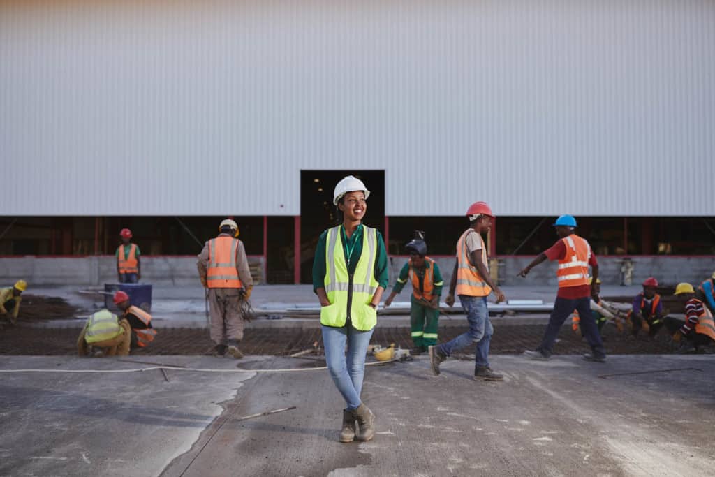 Young woman, in a yellow and gray safety vest, green shirt, blue jeans and white hard hat, is at the plant. She is standing on concrete in front of a large white building. There are workers in orange vests in the background.