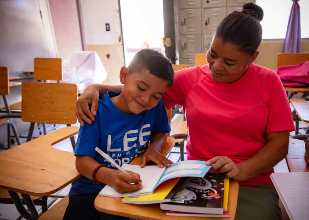 Jose is wearing a blue shirt. He is sitting at a desk and is working on his homework. His grandmother, wearing a red shirt, is sitting next to him and is helping.