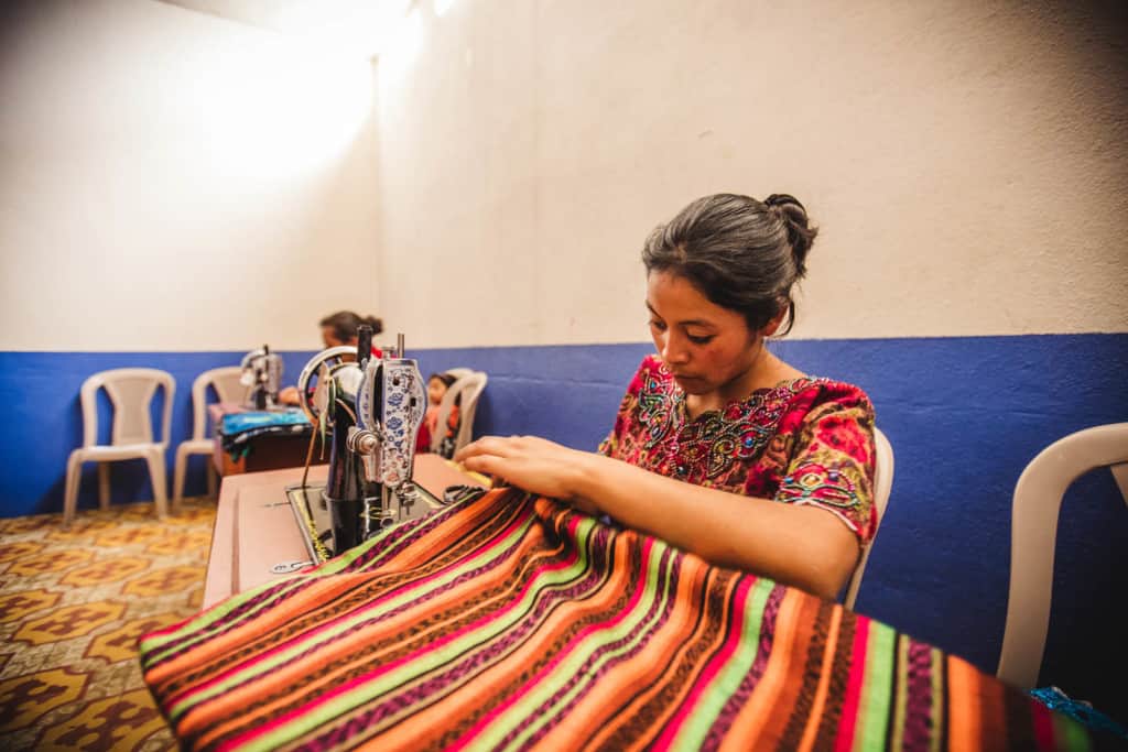 A woman in a red shirt sits in a white chair at a black sewing machine working on a striped piece of fabric that is brown, green, orange and pink. The walls in the room are blue and white.