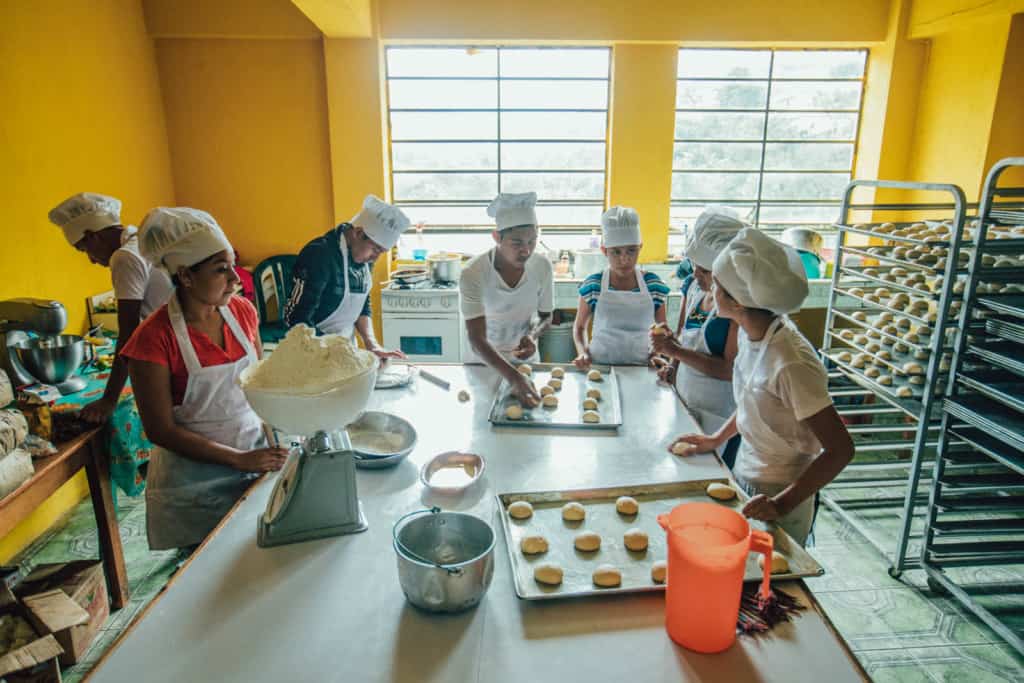 Boys and girls in white aprons and chef hats stand around a white table, counter putting rolls, buns, baked goods onto baking sheets. There is a scale with a bowl of flower on the table. The walls are bright orange, yellow. There are baking sheet racks on one side of the room.