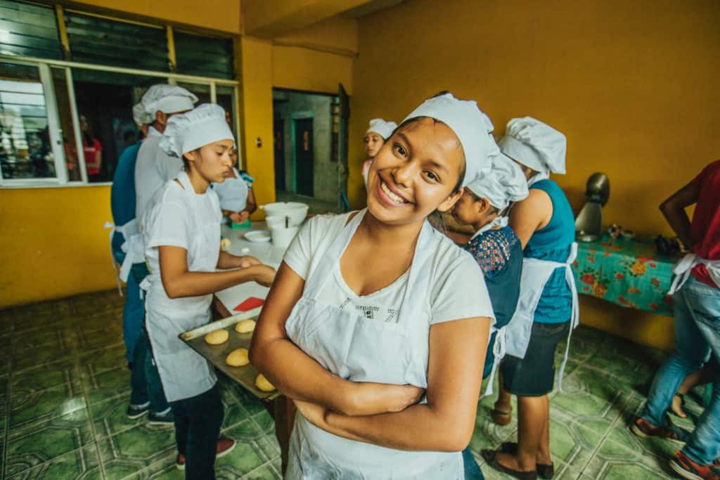 A smiling teen girl in a white apron and chef's hat stands in front of a group of other students who are baking, cooking at a long white table in an orange, yellow room with a green floor.