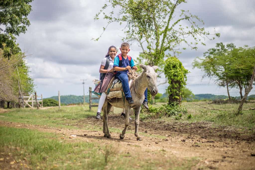 Dilan and Dianis are going to school on their family’s donkey that serves as their transportation. They are wearing their school uniforms and backpacks. They are traveling on a dirt road.
