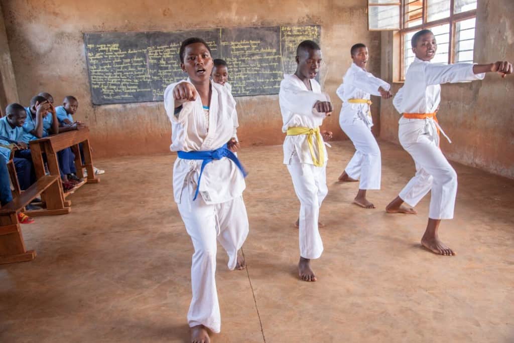 A young woman and four other people in karate gear demonstrates a karate move. She is wearing karate clothing and a blue belt