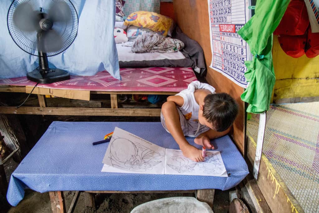 Jasper is sitting on a blue table using his feet to color in a coloring book. Jasper is wearing a white shirt and gray and white striped shorts. There is a calendar on the wall to one side of him. Behind him is his bed and there is a fan blowing on him.