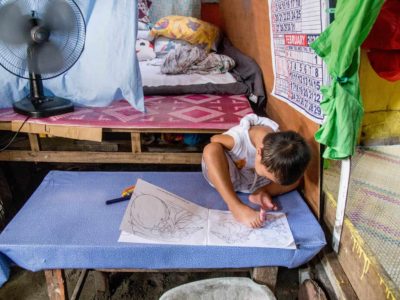 Jasper is sitting on a blue table using his feet to color in a coloring book. Jasper is wearing a white shirt and gray and white striped shorts. There is a calendar on the wall to one side of him. Behind him is his bed and there is a fan blowing on him.