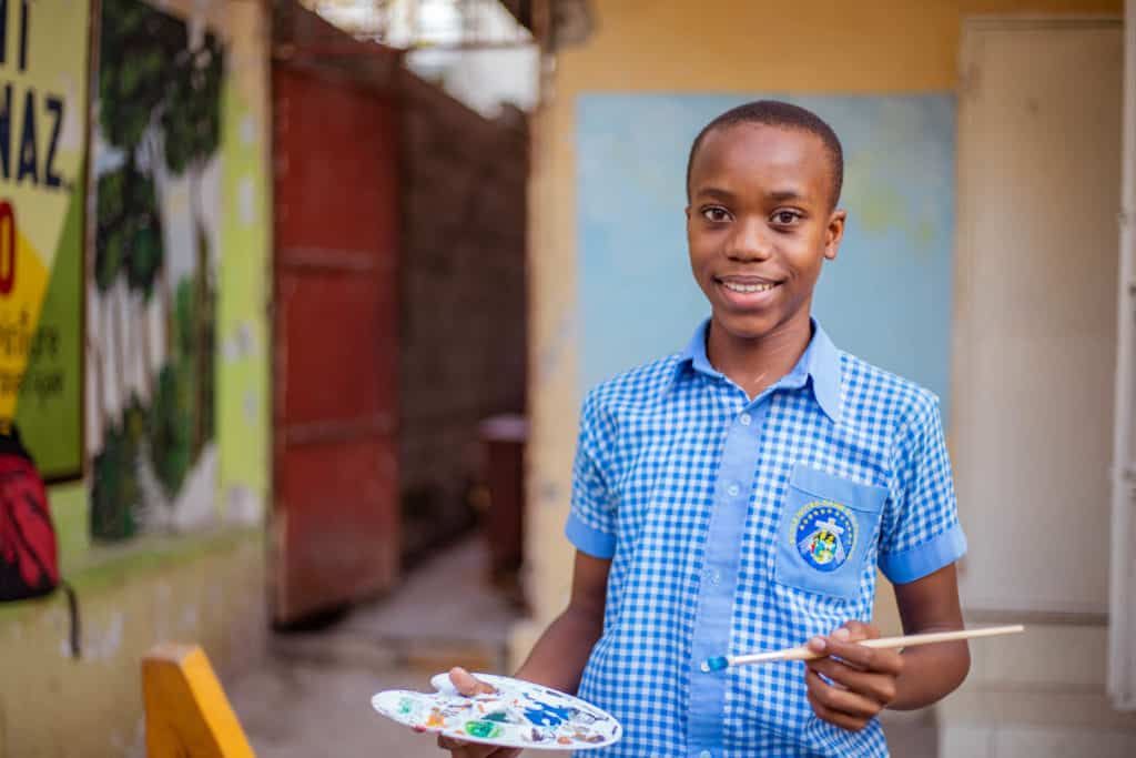 Jetmy has just done a painting of a landscape at his project. He is holding a brush and a plastic palette with orange, blue, green, and red acrylic paints. Jetmy is wearing blue pants and a blue and white checkered shirt.