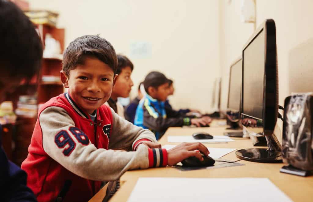 A boy wearing a red and gray jacket with a number patch on the sleeve smiles as he sits in front of a computer. Other children sit at computers nearby.