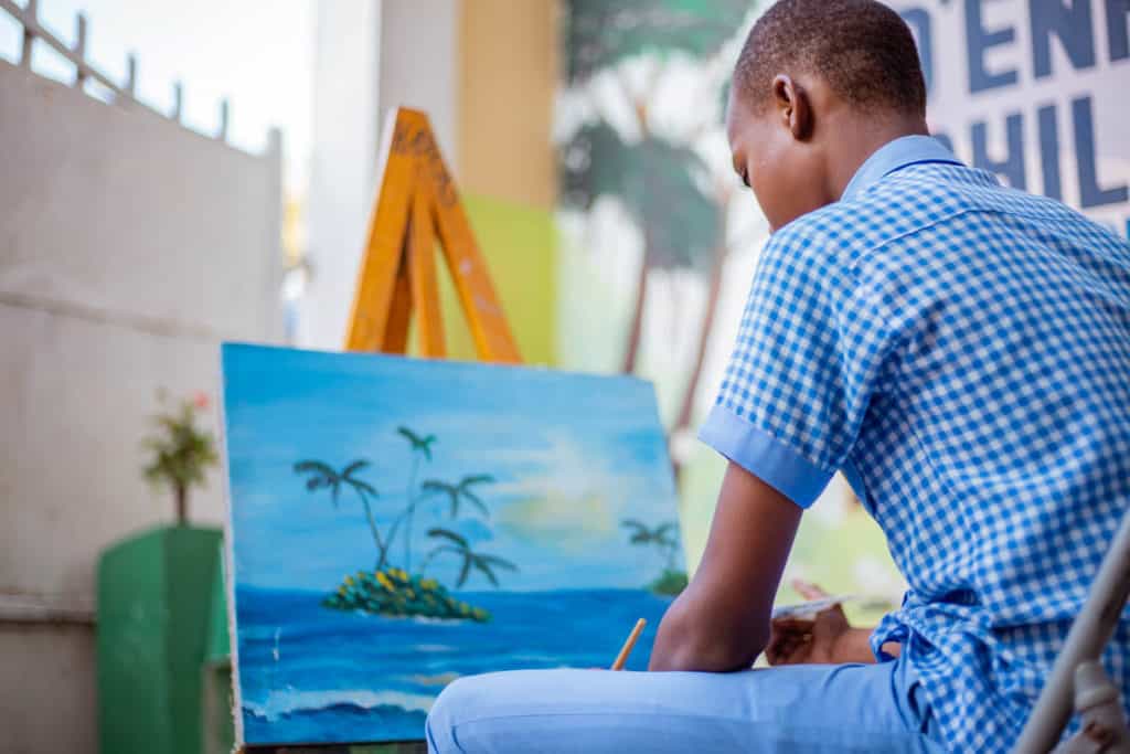 Jetmy is sitting at his project painting a blue sea and sky landscape. Jetmy is wearing his school uniform, blue pants and a blue and white checkered shirt.