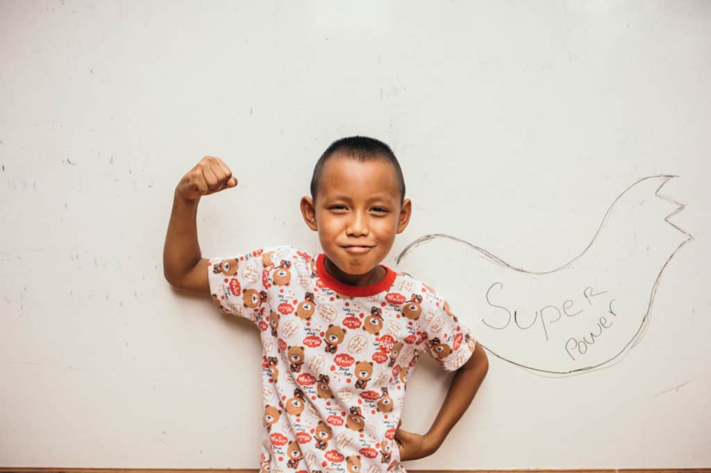 Potikorn is wearing an orange and white print shirt. He is standing in front of a white wall that has a superhero cape drawn on it. He is flexing his muscle.