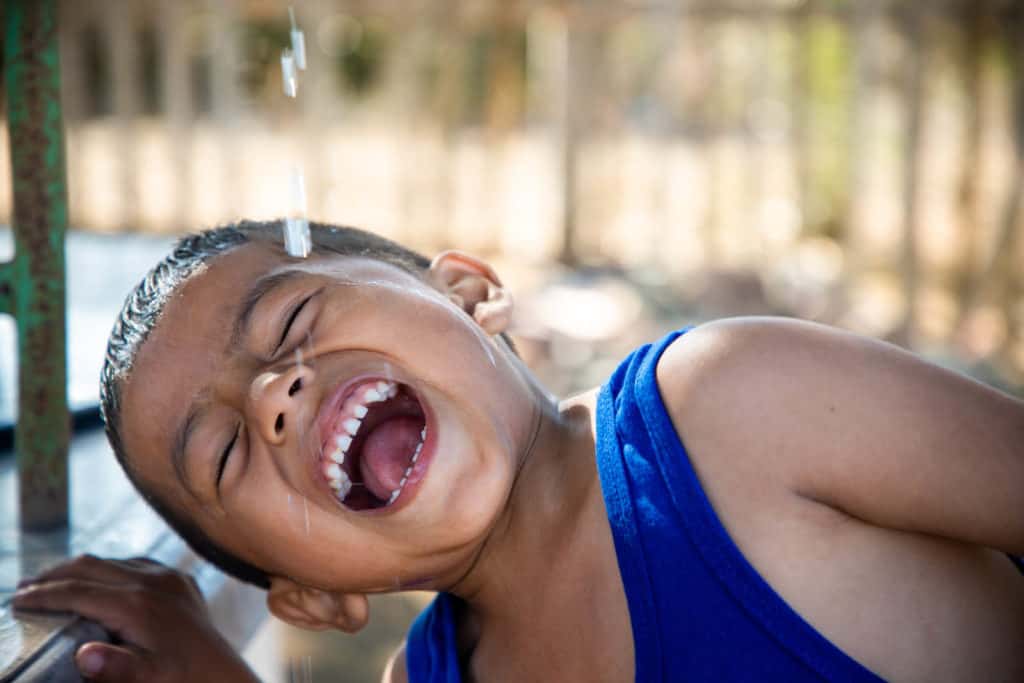 Jimmy is wearing a blue shirt. He is holding his mouth open and is catching water with it.