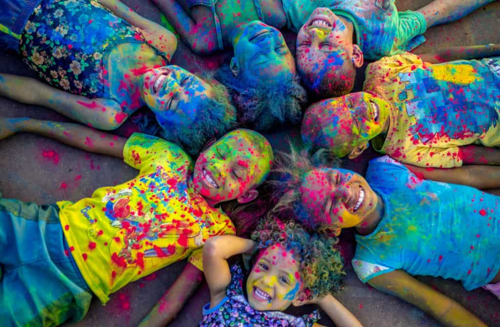 Several children are pictured here, laying in a circle with their heads together. They are covered in colorful powder and are happy and smiling.