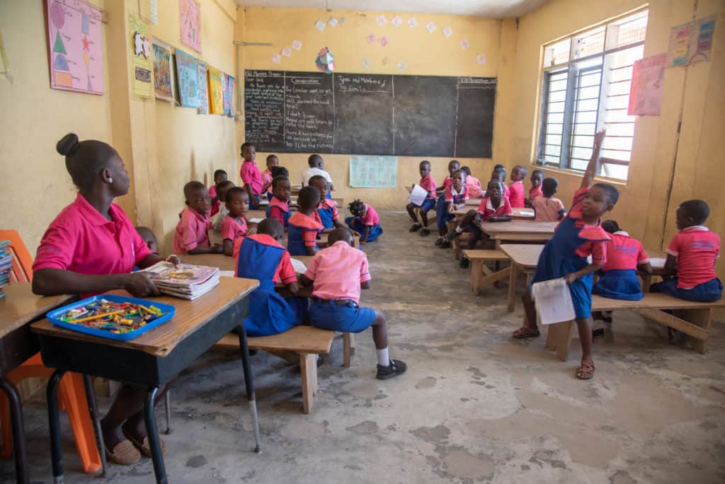 Children, all wearing hot pink and blue uniforms, are in a classroom with yellow walls and a blackboard. The teacher is sitting at a desk holding a set of workbooks.