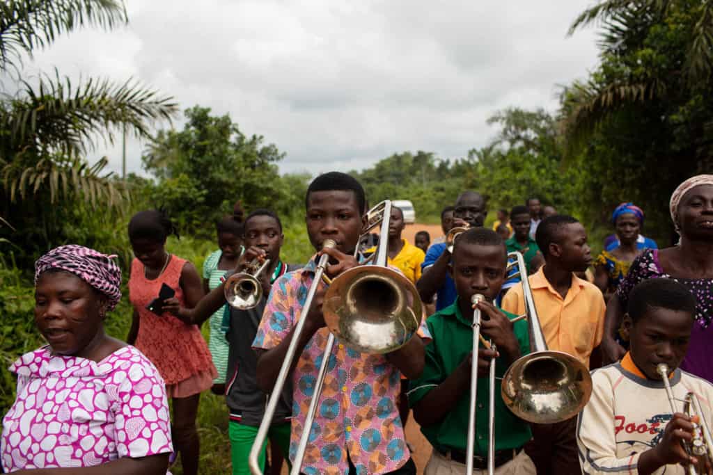 A group of young men are walking down a road bordered by trees playing musical instruments, trombones and trumpets.