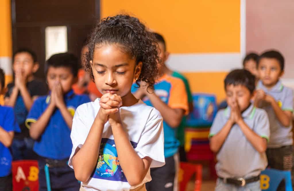 Allison is wearing a white shirt with a design on the front. She is standing in a room with orange walls praying. Her hands are folded in front of her. Other children are behind her praying.