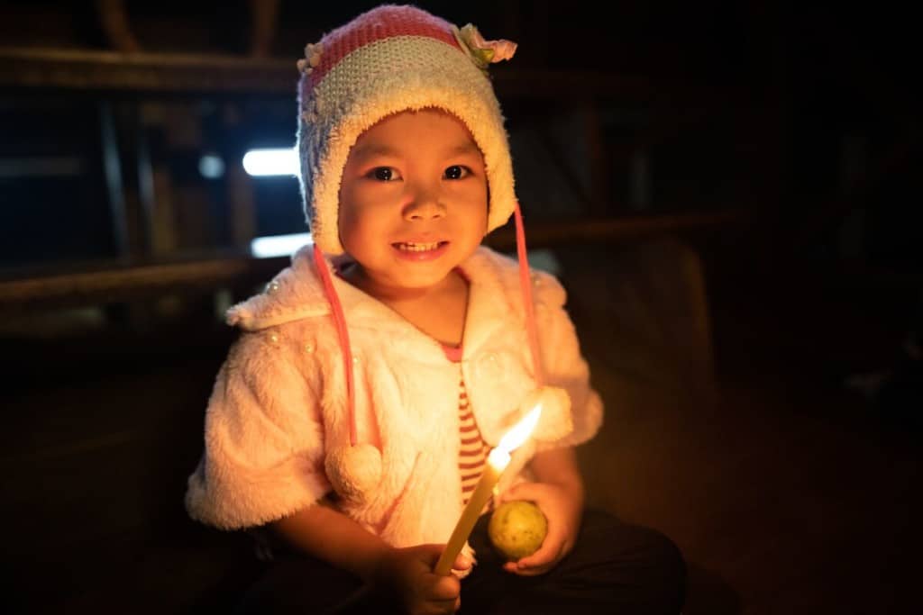 A young child wearing a hat holds a candle in one hand and an orange in the other