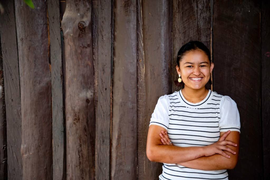 A teen girl in a white and black striped shirt smiles and crosses her arms while standing in front of a wooden fence