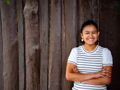 An adolescent woman in a white and black striped shirt smiles and crosses her arms while standing in front of a wooden fence