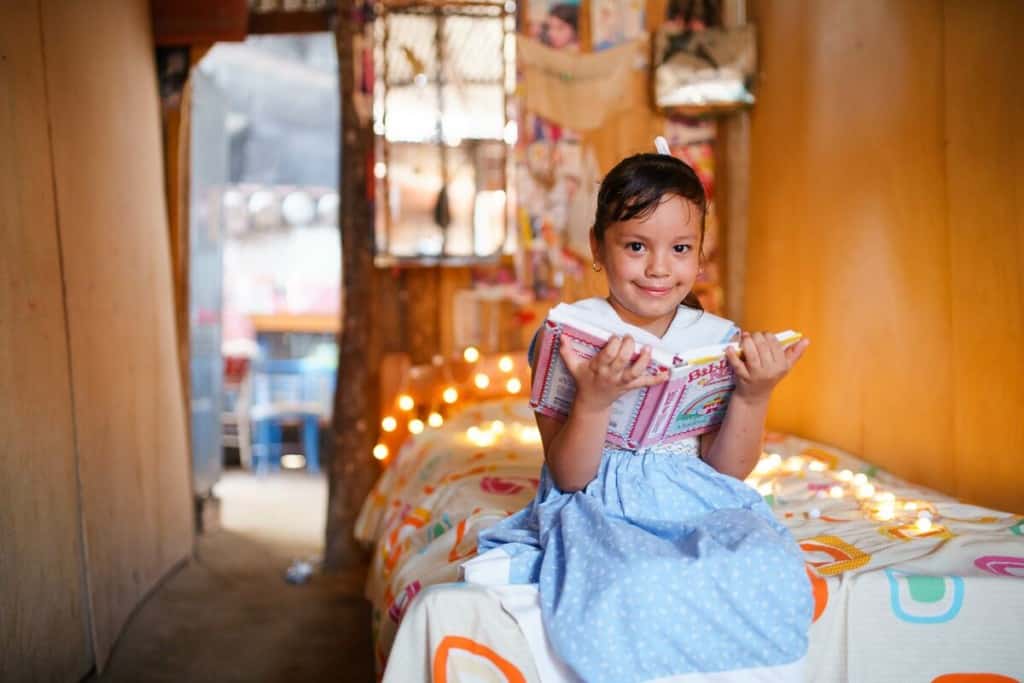 A girl sits on her bed holding an open Bible. There are twinkling lights in the background