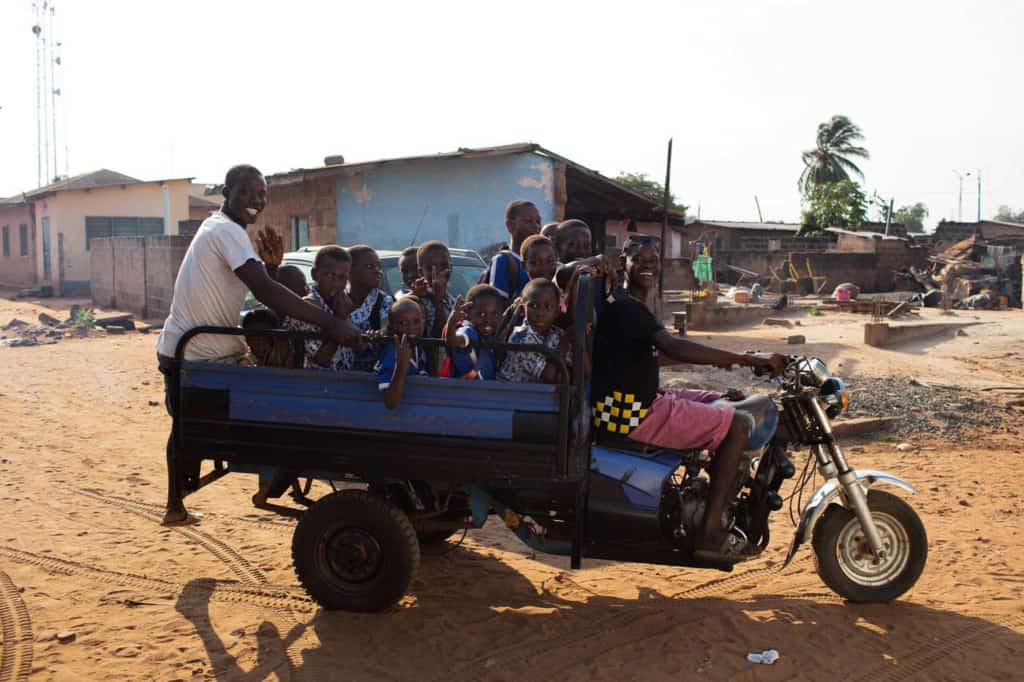 A group of boys and a man in a white shirt are riding in a blue motorcycle truck. There are buildings in the background.
