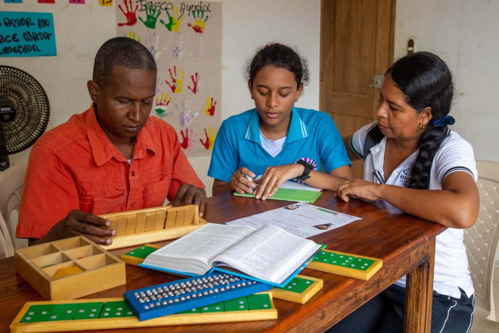 Liseth, wearing a blue shirt, is at the project with her tutor Deysi, wearing a white shirt, and her braille tutor Juan Carlos, wearing a red shirt. Liseth is writing a bible verse in braille.