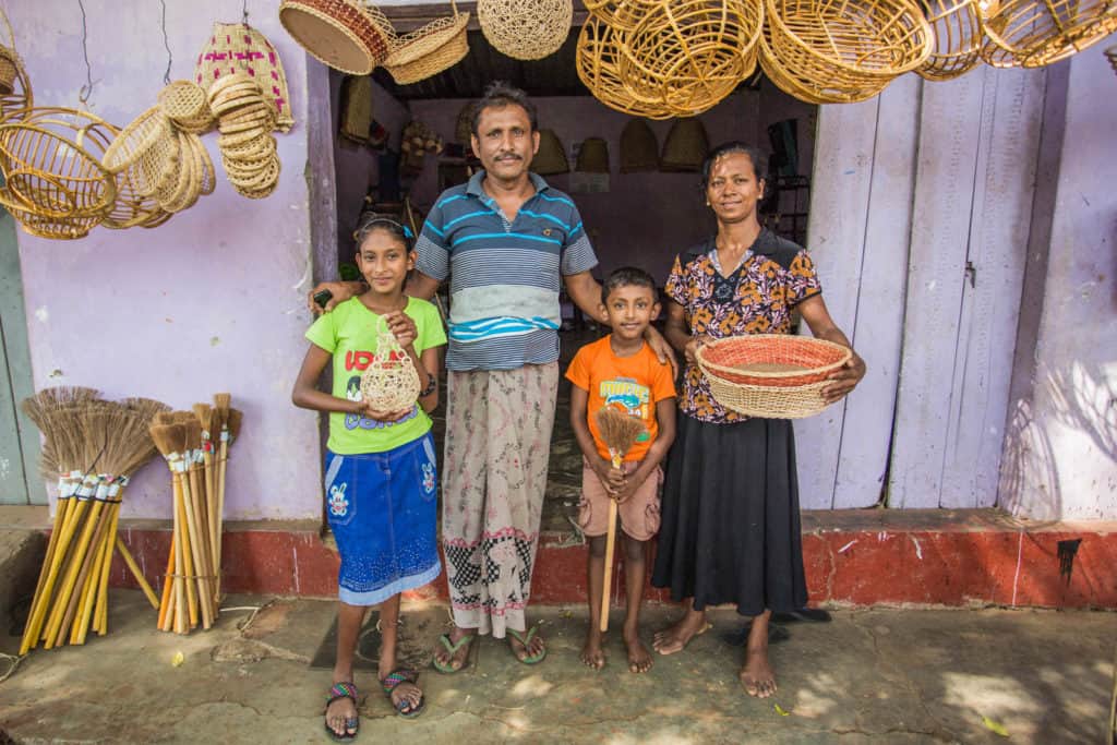 Dilakshi, wearing a blue skirt and a green shirt, is standing with her family in front of their grandmother's home, where they live. There are baskets hanging above them.