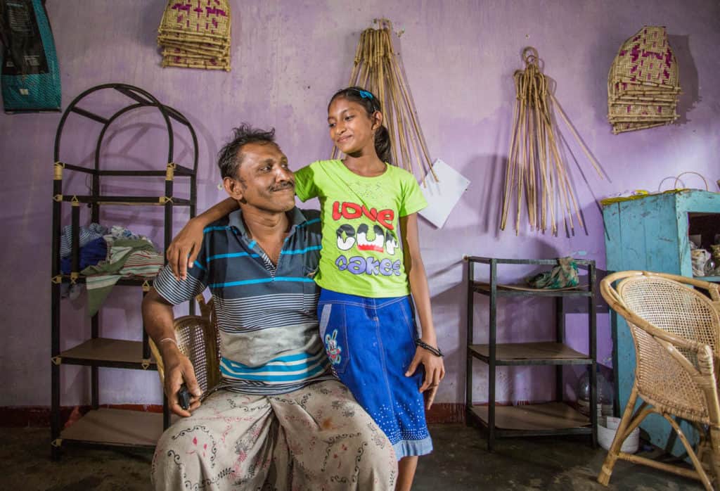 Chamodya Dilakshi, wearing a blue skirt and a green shirt, is with her father, Jayakodi, in his furniture shop that the church helped secure. Jiyakodi is sitting and Chamodya has her arm around him. The wall behind them is purple and there are various pieces of furniture all around them.