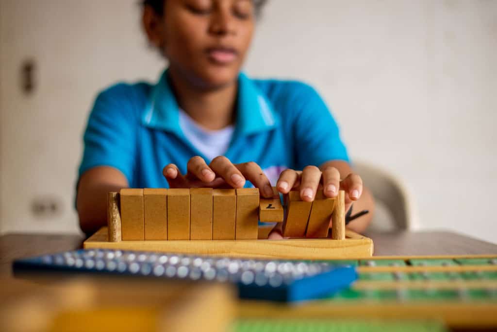 Liseth is sitting at a table and is wearing a blue shirt. She is learning to identify numbers in braille.